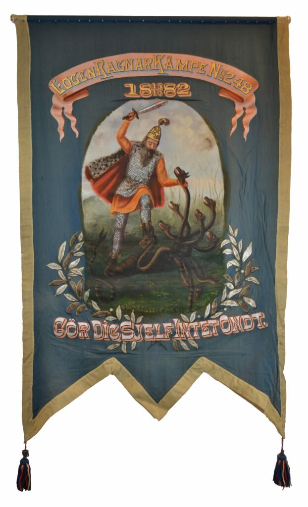 Målat motiv av en riddare som strider mot en månghövdad drake. Riddaren håller ett av drakhuvudena runt halsen och står beredd att hugga av dess huvud. Under målningen en lövkrans och texten "Gör dig sjelf intet ondt". Standarets botten är blå och kanterna är gula.