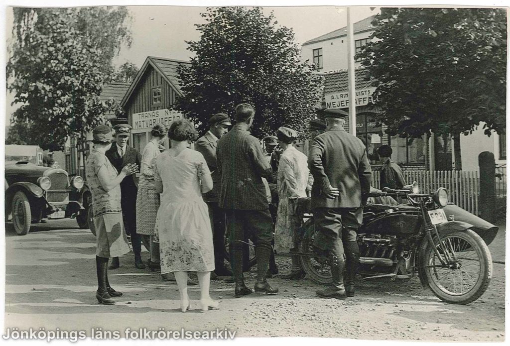 Ett tiotal män och kvinnor samlade runt en motorcykel med sidovagn. I bakgrunden en bil och ett trähus med en skylt som det står Tranås nya matvaruaffär på.