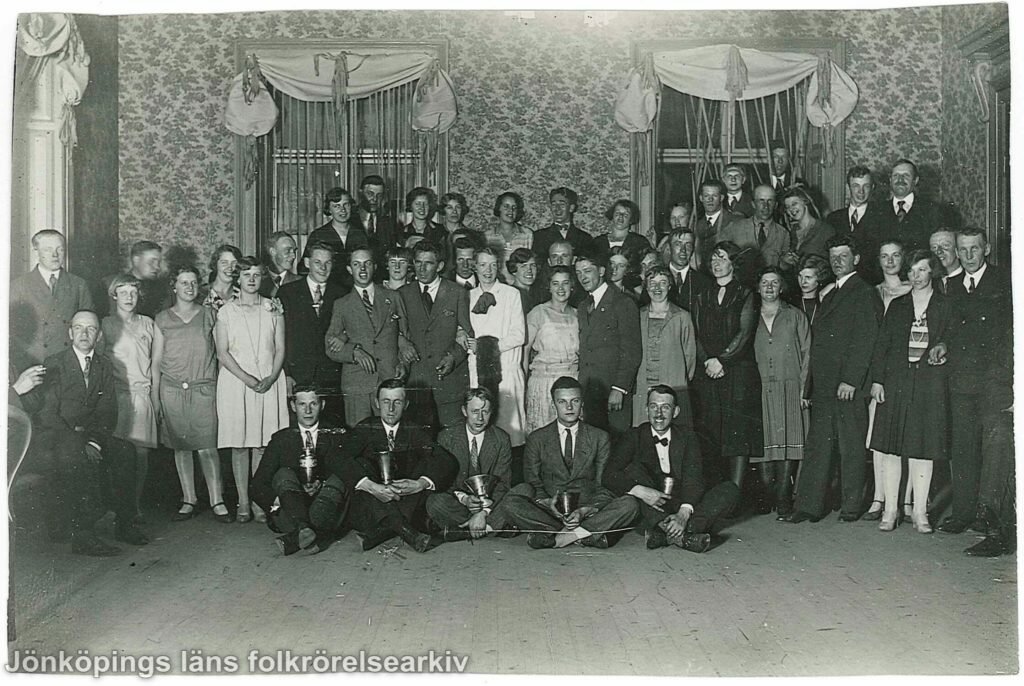 Ett fyrtiotal personer, både män och kvinnor, uppställda för fotografering i en samlingslokal. Längst fram sitter fem män på golvet med benen i kors. I händerna håller de någon slags pokal.