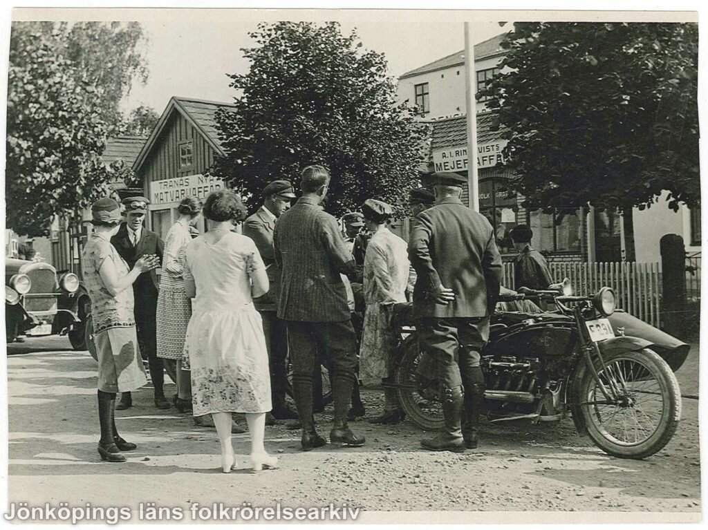 Ett tiotal män och kvinnor samlade runt en motorcykel med sidovagn. I bakgrunden en bil och ett trähus med en skylt som det står Tranås nya matvaruaffär på.