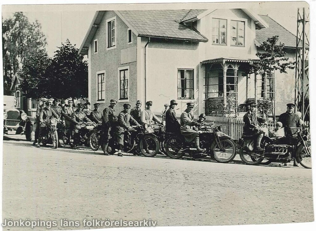 Ett tjugotal personer på motorcyklar framför ett putsat stenhus. En del av motorcyklarna har sidovagnar i vilka barn sitter.