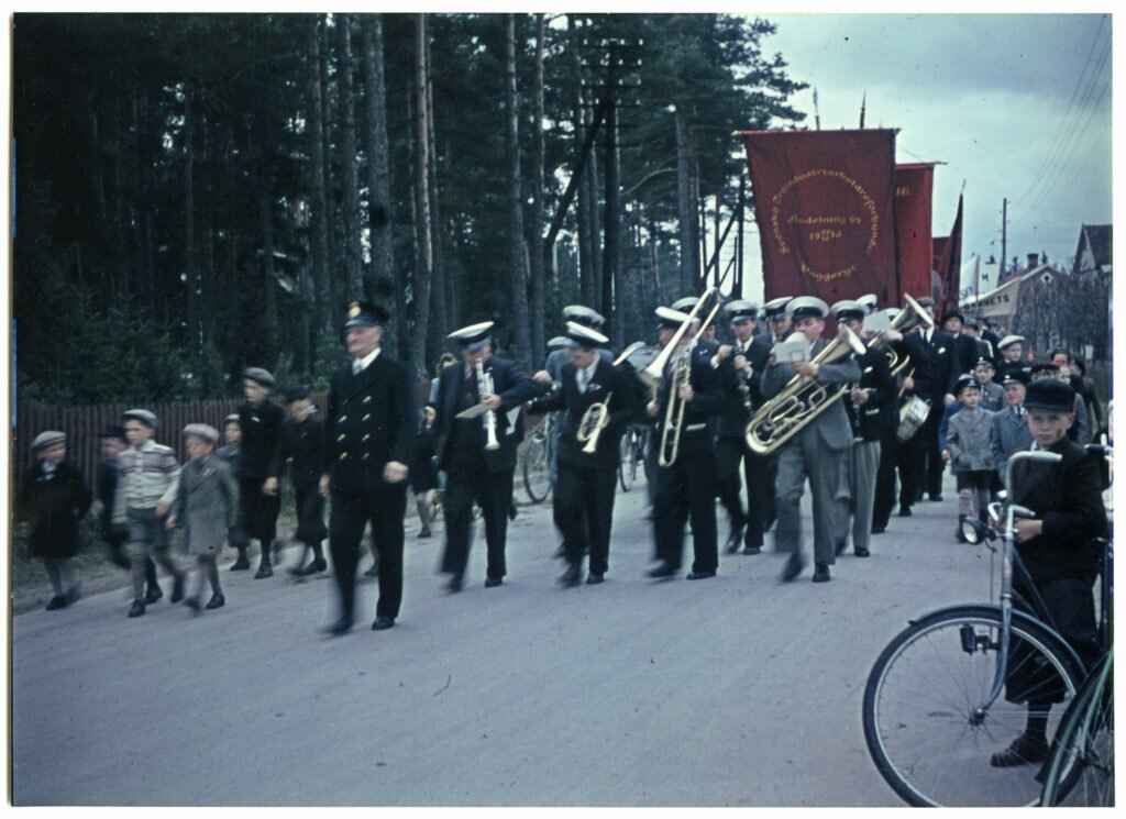 Demonstrationståg med musikanter och fanor går på en väg kantad av åskådare på ena sidan och skog och staket på andra sidan vägen.