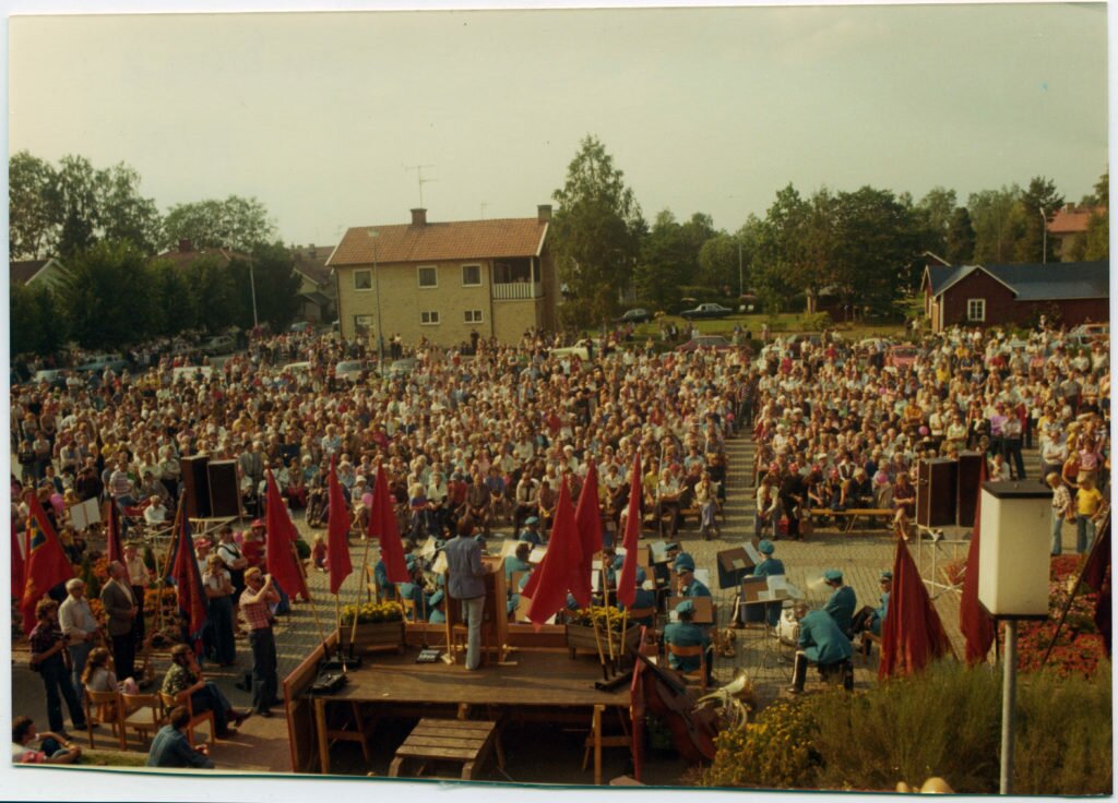 På bilden syns ryggtavlan på en man som står på ett podium och talar inför ett folkhav. Podiet är kantat av röda fanor. I bakgrunden hus, bilar och träd.