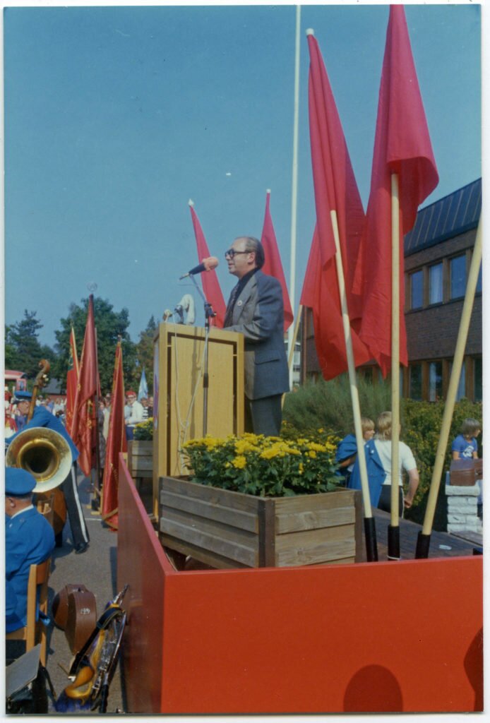 En man står på ett podium kantat av röda fanor och talar inför åhörare.