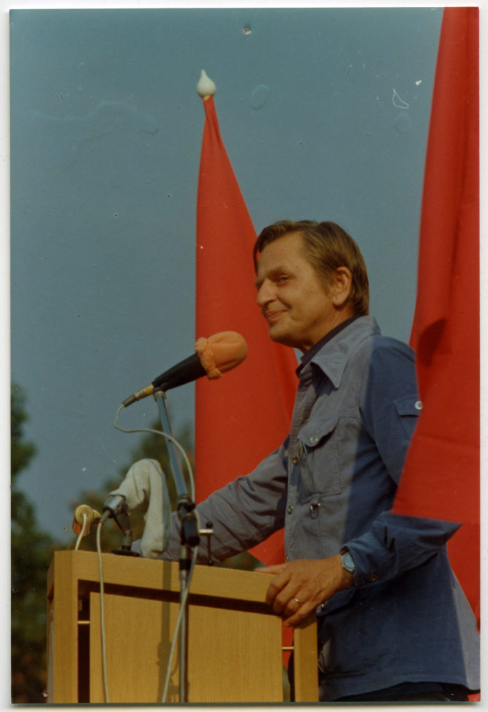En man i halvfigur står vid en mikrofon. Två röda fanor skymtar vid hans sida.