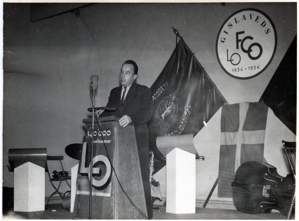 En man står vid en talarstol på en scen med en mikrofon. I bakgrunden En fackfana och en svensk flagga.