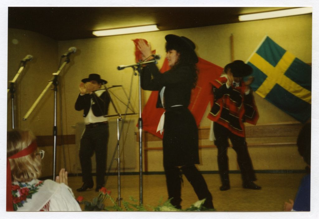 Några män och kvinnor dansar och spelar på en scen. I bakgrunden på väggen hänger en svensk flagga och en LO-fana. Artisternas kläder för tankarna till spansk folkmusik.