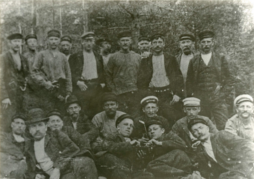 Svartvitt foto på ett tjugotal män i olika åldrar uppställda för foto. I bakgrunden skog.