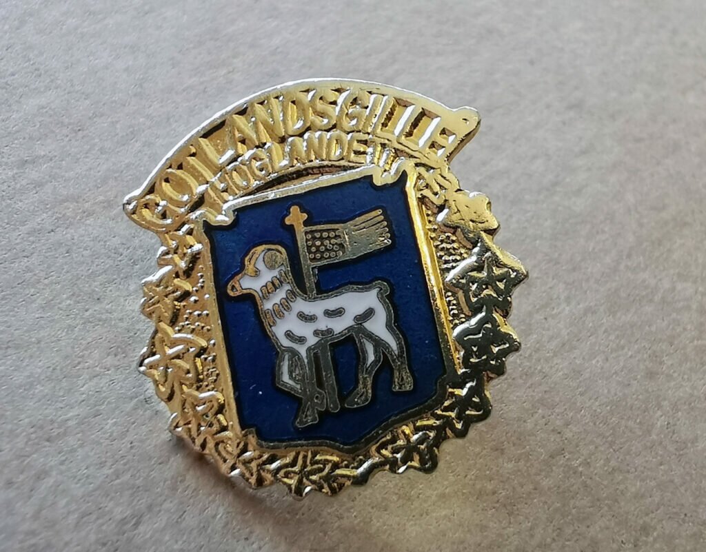 Gyllene nål med får och flagga mot blå botten.