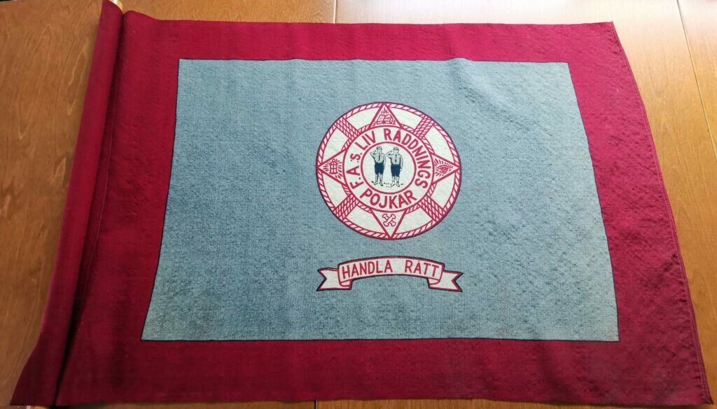 Broderad fana i rött, grått och vitt. I mitten en livboj. Innuti livbojen två scoutpojkar som gör honnör. Broderad banderoll med texten "Handla rätt". 