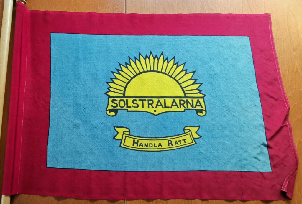Broderad fana i rött, blått och gult. I mitten en halv sol. Under solen texten "Solstrålarna". Längre ner broderad banderoll med texten "Handla rätt". 