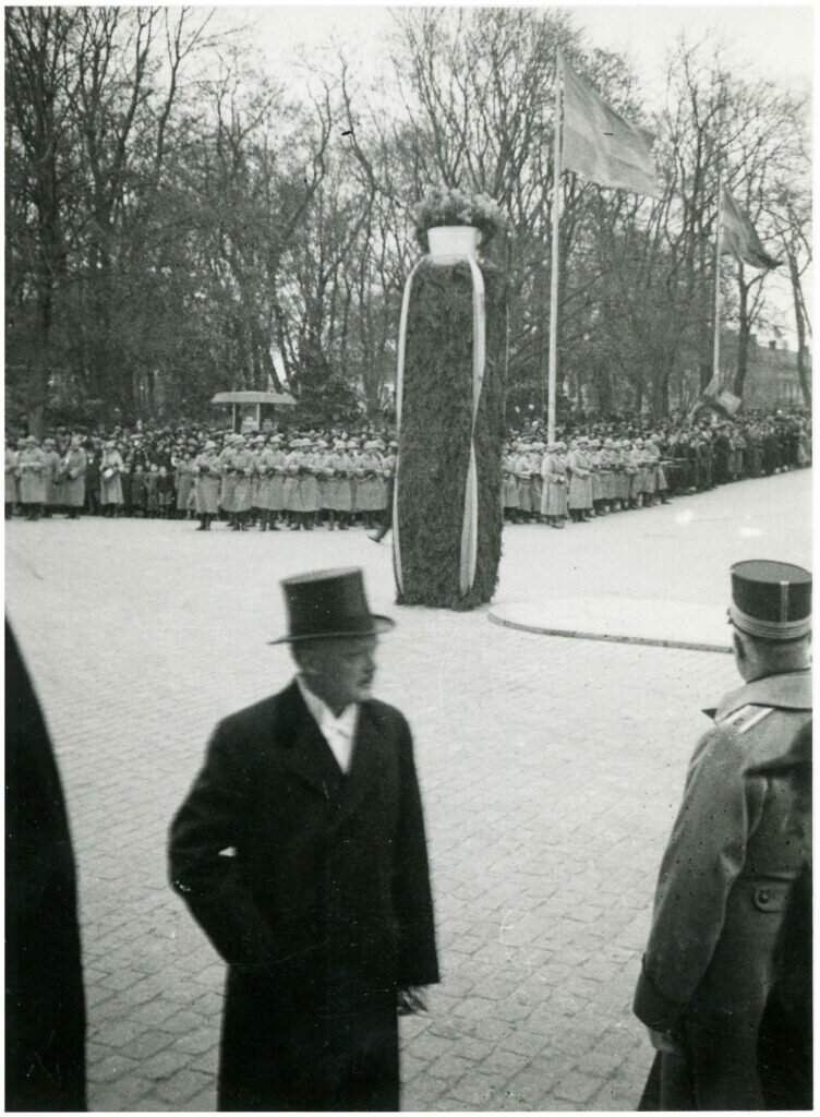 Utomhusfoto. I förgrunden en man i hatt och rock samt ryggen på en militärklädd man. I mitten hög pelare klädd i grönska och blomster. I bakgrunden en stor samling människor.