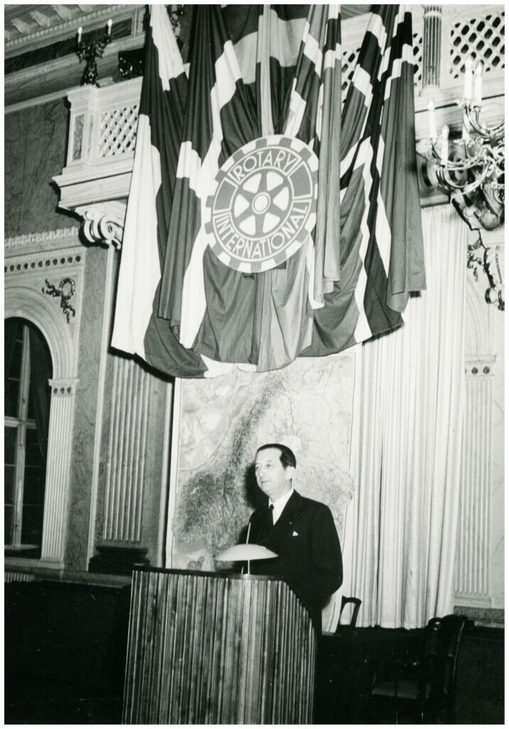 Foto av man vid podium. Ovanför hänger flaggor och vimplar med texten Rotary International. 