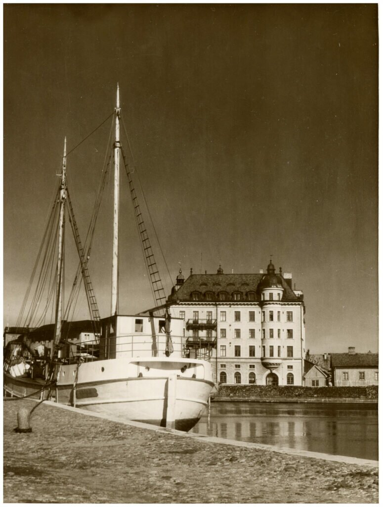 Foto av båt vid kajplats. I bakgrunden stenhus. 