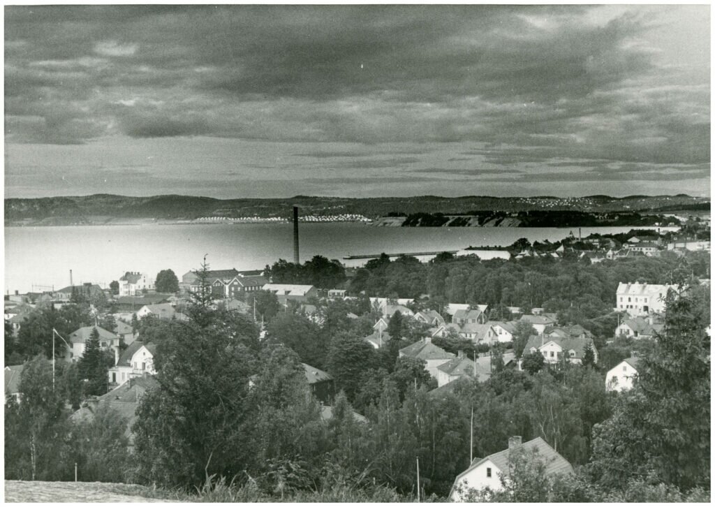 Vy över stad vid vatten. I mitten tornar en skorsten upp sig. Längre bort syns sandbankar som går ner i vattnet. 