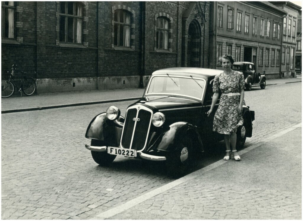 Foto av kvinna i klänning stående vid en bil. Förmodligen 1940- eller 1950-tal. Kullerstensgata. I bakgrunden stenhus. 