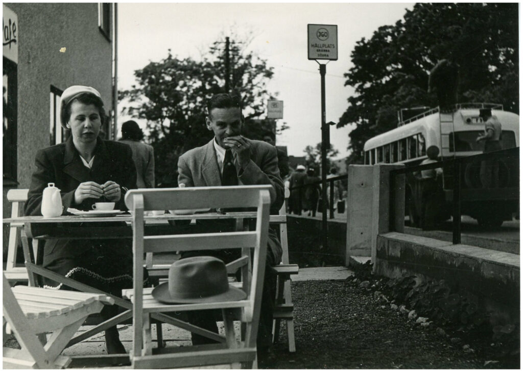 En kvinna och en man sitter vid ett bord och fikar. I bakgrunden en hållplats med en buss. 