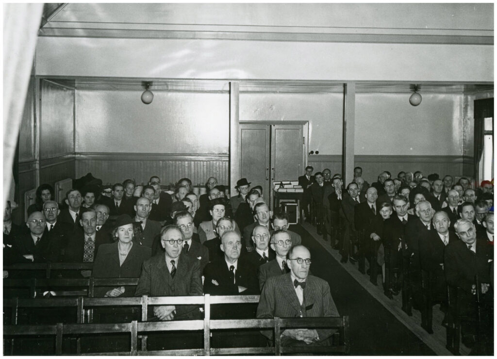 Foto av människor sittandes i bänkar samlade för möte i sal. 